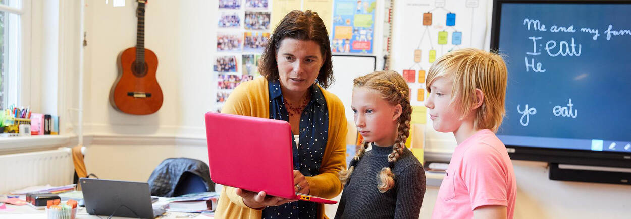 Leerkracht geeft met laptop uitleg aan 2 leerlingen