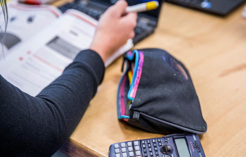 Meisje maakt aan tafel opdracht in werkboek, rekenmachine en laptop liggen op tafel